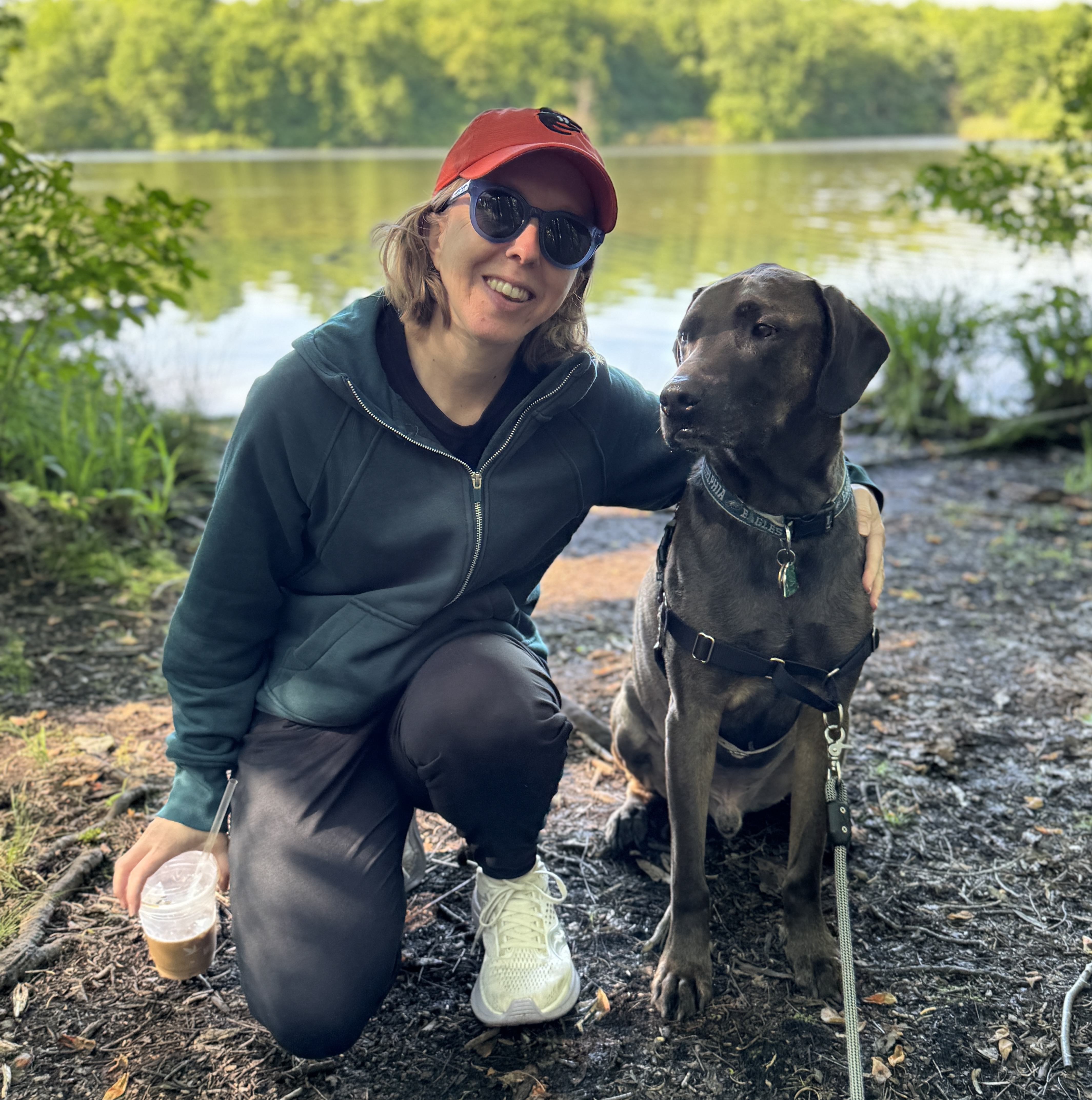 A woman kneeling down to take a picture next to her dog at the edge of a lake.